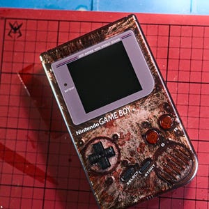 Könnte beinhalten: Eine individuell lackierte Nintendo Game Boy-Handheld-Konsole. Das Gerät hat eine braune, strukturierte Oberfläche mit roten und schwarzen Akzenten. Der Bildschirm und die Tasten sind sichtbar, mit dem Text "Nintendo GAME BOY" und "DOT MATRIX WITH STEREO SOUND".