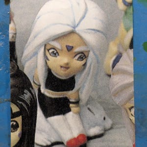 Puede incluir: Una muñeca de pelo blanco con un atuendo azul y morado y zapatos rojos. La muñeca tiene una capa con capucha azul claro y blanca. La muñeca está sentada sobre una superficie blanca.