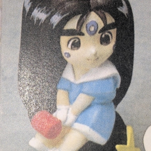 May include: A figurine of a cartoon character with black hair, wearing a blue dress with white trim. The character has a red object in their hands and a yellow star next to them.