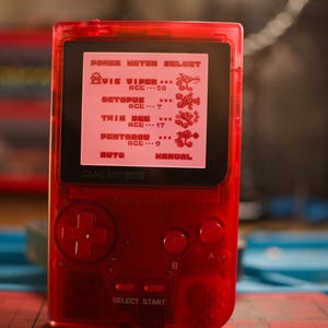 May include: A transparent red Game Boy Pocket handheld console, displaying a game screen with pixelated graphics. The screen shows text and character sprites. The console has a directional pad, buttons, and the text "SELECT START".