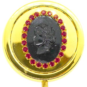 May include: A gold-colored compact mirror with a black cameo portrait surrounded by red gemstones. The compact is round and has a small clasp. The cameo features a profile of a woman.