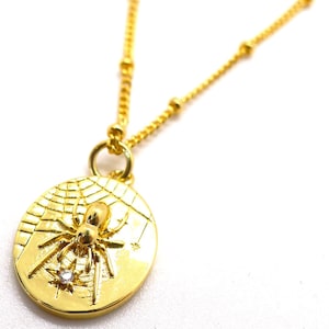 May include: Gold-toned necklace featuring a pendant with a spider and web design. The pendant has a small, sparkling accent. The chain is delicate with small, round beads. The necklace is set against a white background.