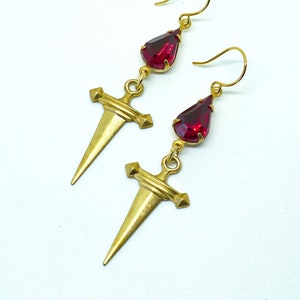 May include: Gold dagger earrings with red teardrop-shaped crystals. The daggers have a pointed tip and a cross-shaped top.