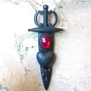 Puede incluir: Un collar con colgante de metal oscuro con un diseño de daga con un corazón y una gema ovalada roja. La daga tiene una empuñadura decorativa y está suspendida de una cadena plateada.