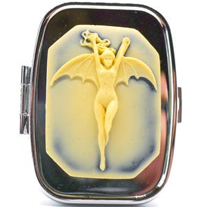May include: A silver-toned compact mirror with a cream-colored cameo featuring a winged figure with outstretched arms. The cameo is set against a light blue background within the rectangular mirror.