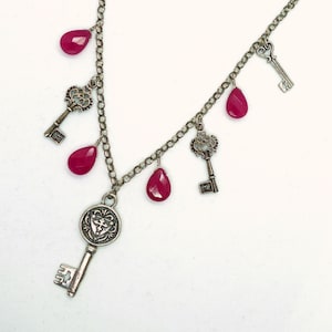Puede incluir: Collar plateado con llaves decorativas y gemas rojas en forma de lágrima. El collar tiene un diseño de cadena con múltiples llaves y gemas colgando de él. Las llaves tienen detalles intrincados y las gemas son de color rojo vibrante.