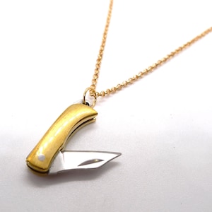 Puede incluir: Un collar con colgante dorado con un dije de cuchillo en miniatura. El cuchillo tiene una hoja plateada y un mango curvo en tono dorado. La cadena del collar es de color dorado delicado.