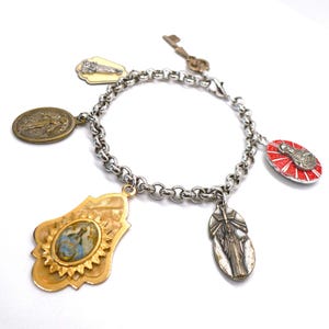 Könnte beinhalten: Ein silberfarbenes Charm-Armband mit verschiedenen religiösen Anhängern. Zu den Anhängern gehören ein goldfarbenes Oval mit einem blau-weißen Bild, ein rotes Oval mit einem silbernen Bild und ein schlüsselförmiger Anhänger. Weißer Hintergrund.