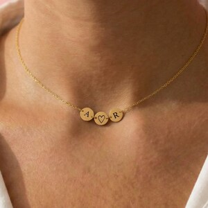 Pode incluir: Um colar dourado com três pequenos pingentes redondos. Os pingentes são gravados com as letras "A", "R" e um símbolo de coração. O colar é usado sobre uma camisa branca.