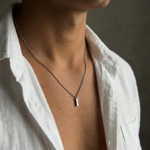 Peut inclure: Un collier pendentif argenté avec un charm rectangulaire. Le collier est suspendu à une chaîne argentée. La personne porte une chemise blanche boutonnée.