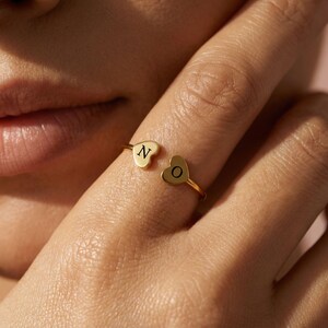 Peut inclure: Une bague dorée avec deux extrémités en forme de cœur, chacune portant une initiale. Un cœur affiche la lettre "N", l'autre la lettre "O". La bague est portée au doigt, présentant un bijou délicat et personnalisé.