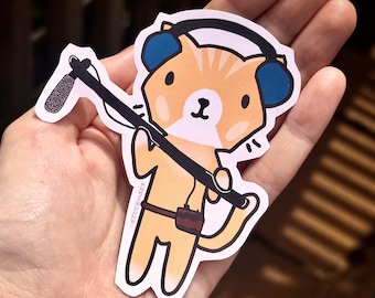Puede incluir: Una pegatina con un gato de dibujos animados con pelaje naranja, que lleva auriculares azules y sostiene una pértiga de micrófono. El gato tiene un cinturón marrón pequeño con una bolsa. La pegatina tiene un borde blanco y el texto "STUCK ON SET".