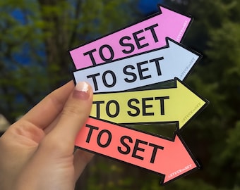 Puede incluir: Cuatro pegatinas de flecha de colores con el texto "To Set" en letras negras. Las pegatinas están superpuestas, con la pegatina rosa en la parte superior y la pegatina naranja en la parte inferior. Las pegatinas se sostienen en una mano.