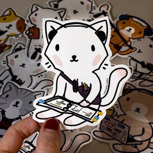 May include: White cat sticker with black headphones, a purple bag, and a notebook. The cat has pink cheeks and is surrounded by other cat stickers. The sticker has the text "STUCK ON SET".