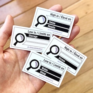 May include: Four rectangular white stickers with black borders and text. Each features a magnifying glass icon and labels like High in / Élevé en Takes, Resets, Low in / Limité en Storage, High in / Élevé en Stress, and Low in / Limité en Water, Sleep with @StuckOnSet.