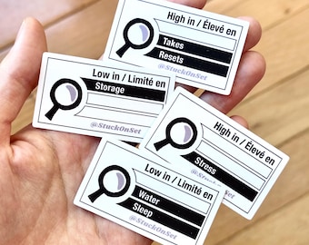 Puede incluir: Cuatro pegatinas rectangulares blancas con bordes y texto en negro. Cada una presenta un icono de lupa y etiquetas como High in / Élevé en Takes, Resets, Low in / Limité en Storage, High in / Élevé en Stress, y Low in / Limité en Water, Sleep con @StuckOnSet.