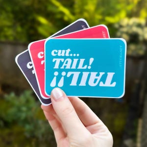 May include: Three stickers with the text "cut... TAIL! i'i TAIL" in white on a turquoise background. The stickers are layered on top of each other, with the turquoise sticker on top.