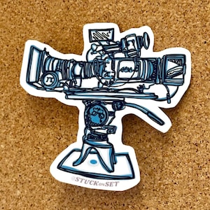 Puede incluir: Una pegatina con una ilustración detallada en azul y blanco de una cámara de televisión vintage sobre un trípode. La pegatina tiene un borde blanco e incluye el texto "@STUCKONSET". La cámara tiene una lente grande y varios diales y pantallas.