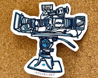 Puede incluir: Una pegatina con una ilustración detallada en azul y blanco de una cámara de televisión vintage sobre un trípode. La pegatina tiene un borde blanco e incluye el texto "@STUCKONSET". La cámara tiene una lente grande y varios diales y pantallas.