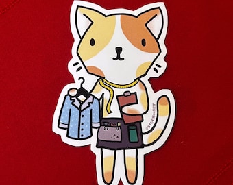 Puede incluir: Una pegatina con un gato de dibujos animados vestido de sastre, sosteniendo una camisa de pijama azul en una percha y una carpeta. El gato tiene pelaje naranja y blanco y lleva una cinta métrica alrededor del cuello. La pegatina tiene el texto "STUCK ON SET".