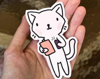 Puede incluir: Una pegatina de gato blanco con una carpeta rosa y un teléfono morado. El gato tiene mejillas rosadas y ojos negros. La pegatina se sostiene en una mano.