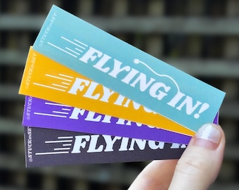 Puede incluir: Cuatro pegatinas con el texto "FLYING IN!" en letras blancas sobre fondos de colores diferentes. Las pegatinas están apiladas una encima de la otra, con la pegatina azul en la parte superior y la pegatina negra en la parte inferior. Las pegatinas son todas del mismo tamaño y forma, y tienen un diseño simple.