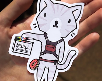 Puede incluir: Una pegatina con un gato de dibujos animados sosteniendo una claqueta de cine. El gato es de color gris claro con rayas grises más oscuras y lleva un arnés granate. La claqueta tiene el texto "A02 | 48 | 1 Stuck On Set". Una bobina de película arcoíris cuelga de la cintura del gato.