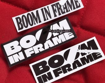Puede incluir: Tres pegatinas en blanco y negro con el texto "BOOM IN FRAME" en una fuente gráfica en negrita. Las pegatinas están sobre una superficie acolchada roja.