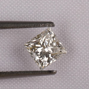 Könnte beinhalten: Ein quadratisch geschliffener Diamant, der von einer Pinzette gehalten wird. Der Diamant hat eine brillante, facettenreiche Oberfläche, die das Licht reflektiert und einen funkelnden Effekt erzeugt. Der Edelstein hat eine helle, cremeweiße Farbe. Die Pinzette ist dunkelgrau.