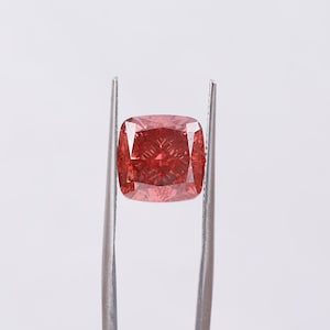 Op de afbeelding: Een roodroze edelsteen met kussensnede wordt door een zilveren pincet tegen een witte achtergrond gehouden. De vierkante steen heeft afgeronde hoeken en interne facetten die het licht reflecteren.