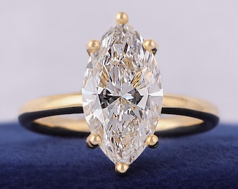 May include: A marquise-cut diamond engagement ring with a gold band. The diamond is set with six prongs and is the focal point of the ring. The band is a simple, polished gold. The ring is displayed on a blue surface.