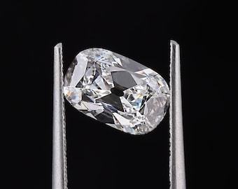 May include: A clear, cushion-cut diamond held by silver tweezers against a black background. The diamond has many facets, reflecting light and creating a brilliant sparkle. The shape is elongated, with rounded corners.