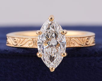 May include: A gold engagement ring with a marquise-cut diamond center stone. The band features an ornate, engraved design. The ring is set against a dark blue background, highlighting the sparkle of the diamond.