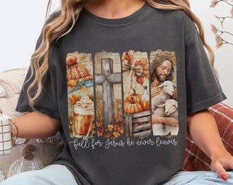 May include: Dark grey t-shirt featuring a fall-themed design. The graphic includes a cross, pumpkins, a latte, and Jesus with lambs. The text "fall for Jesus he never leaves" is printed below the graphic.