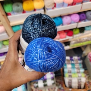 May include: Two round skeins of yarn held together. The top skein is a dark metallic navy blue, and the bottom skein is a vibrant metallic royal blue. Both skeins feature a shiny, reflective thread woven throughout the soft fiber texture.