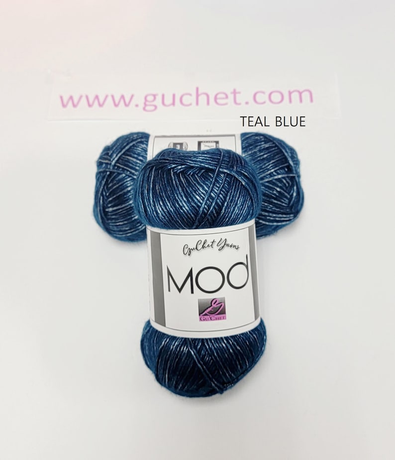 May include: Three skeins of teal blue yarn with a metallic sheen. The central label features the text Guchet Yarns MOD and a logo. The background includes the text www.guchet.com and the color name TEAL BLUE. The yarn has a twisted texture.