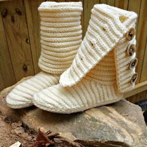 May include: A pair of cream-coloured crocheted slipper boots with a fold-over cuff and button accents. The boots are sitting on a rock.