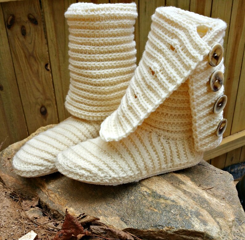 May include: A pair of cream-coloured crocheted slipper boots with a fold-over cuff and button accents. The boots are sitting on a rock.