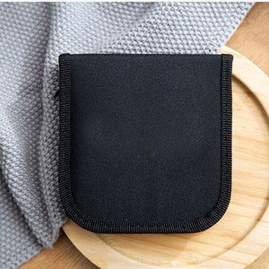 May include: A black zippered pouch with a smooth, textured surface. The pouch is sitting on a wooden surface.