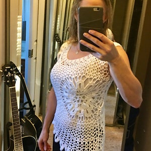 May include: A white crochet top with a scalloped hem. The top is sleeveless and has a loose fit. The woman is wearing black pants.