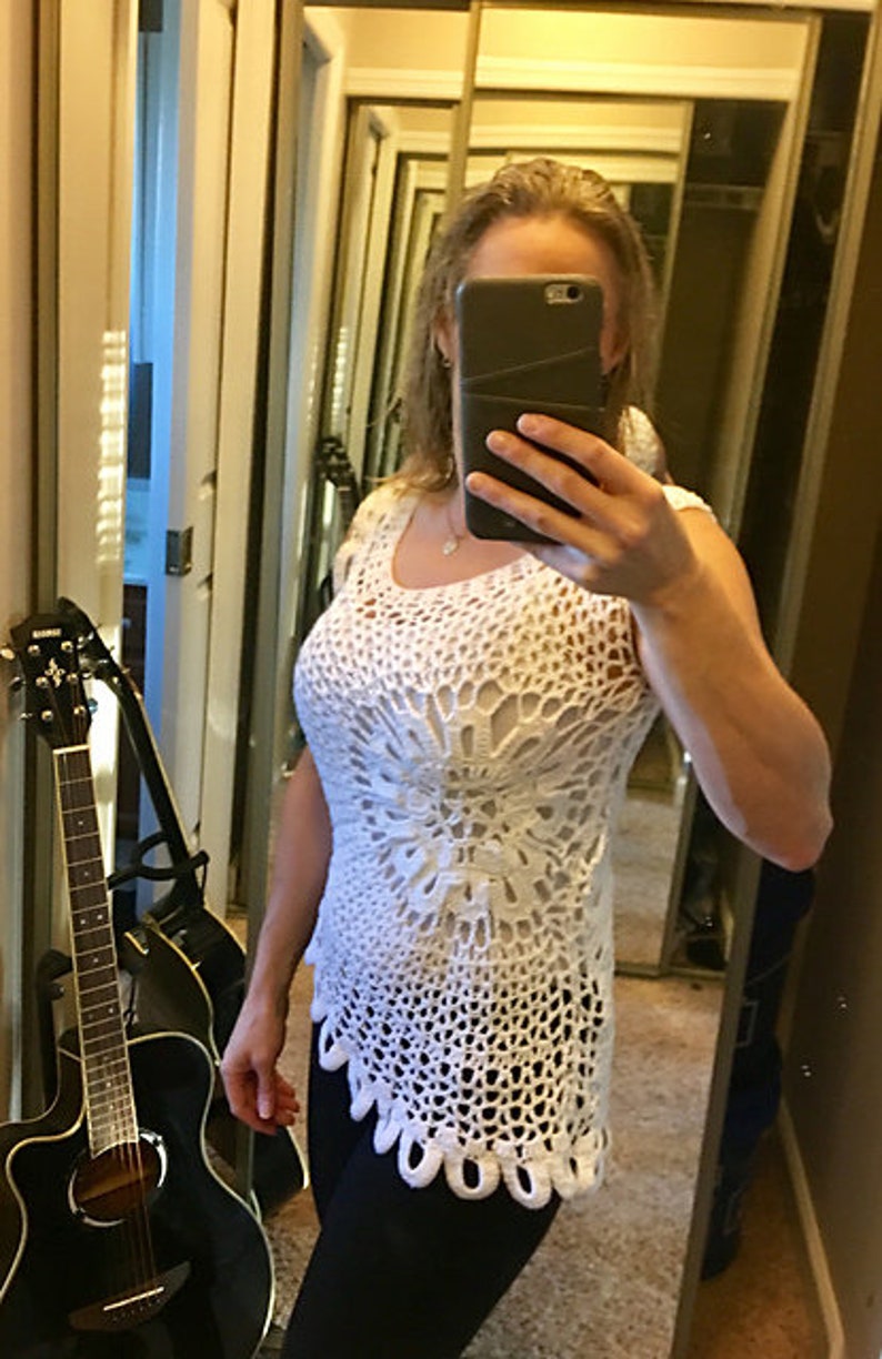 May include: A white crochet top with a scalloped hem. The top is sleeveless and has a loose fit. The woman is wearing black pants.