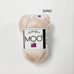 May include: A ball of light beige yarn labeled SAND. The white paper label features the text Crochet Yarns MOD with a pink logo. The yarn has a soft, slightly fuzzy texture and a pale, neutral color tone, presented as a single skein of craft material.