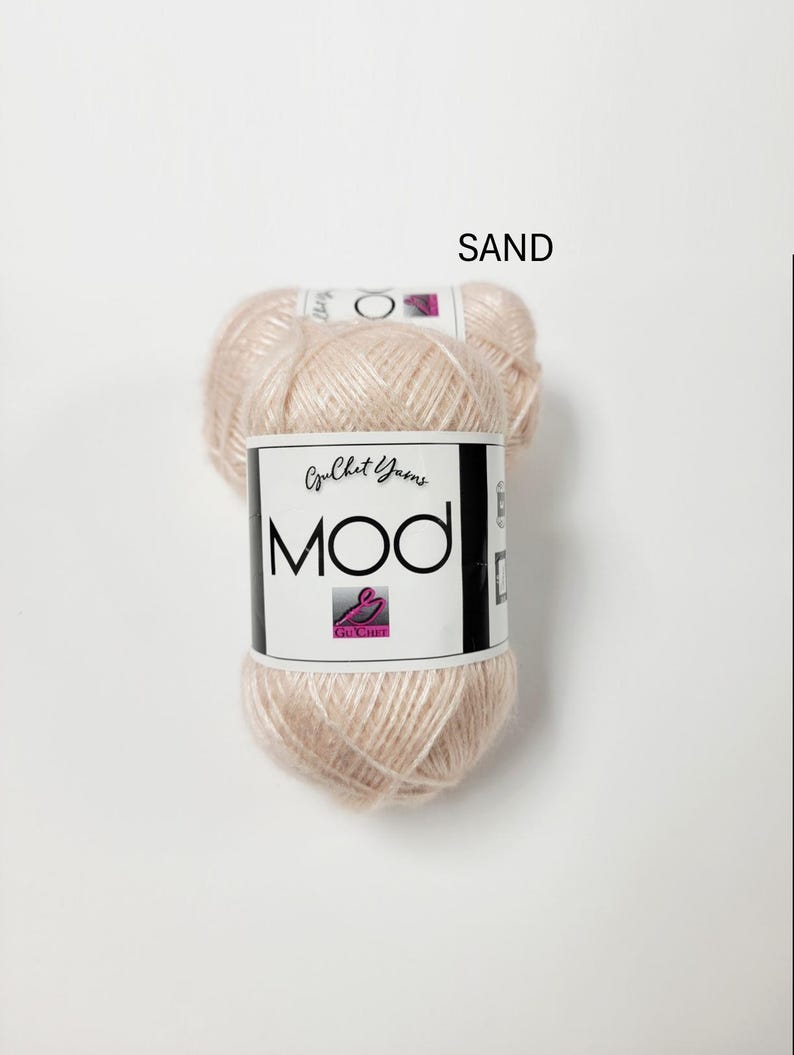 May include: A ball of light beige yarn labeled SAND. The white paper label features the text Crochet Yarns MOD with a pink logo. The yarn has a soft, slightly fuzzy texture and a pale, neutral color tone, presented as a single skein of craft material.