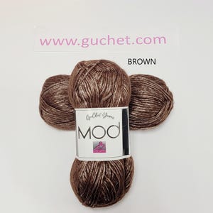 May include: A skein of brown yarn with a metallic sheen, featuring a white paper label that reads Guchet Yarns MOD. The text www.guchet.com and the word BROWN are printed above the yarn. The yarn is wound into a compact, rounded shape.
