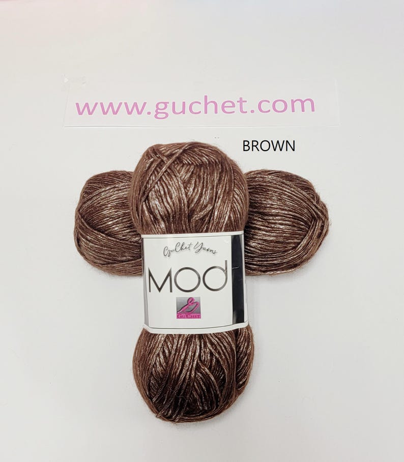 May include: A skein of brown yarn with a metallic sheen, featuring a white paper label that reads Guchet Yarns MOD. The text www.guchet.com and the word BROWN are printed above the yarn. The yarn is wound into a compact, rounded shape.