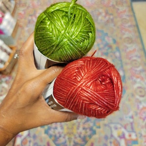 May include: Two round skeins of yarn, one in a vibrant metallic green and the other in a deep metallic red. Each skein is wrapped with a white paper label around the center, showcasing a shiny, textured fiber composition with a distinct sheen.