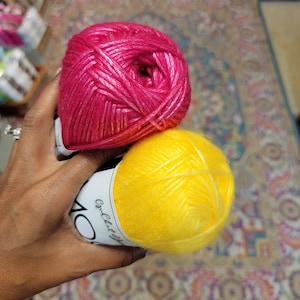 May include: Two skeins of yarn, one in a vibrant magenta pink and the other in a bright sunshine yellow. Each skein features a white paper label with the text Crochet Up, showcasing a soft, fuzzy texture with a subtle sheen throughout the fibers.