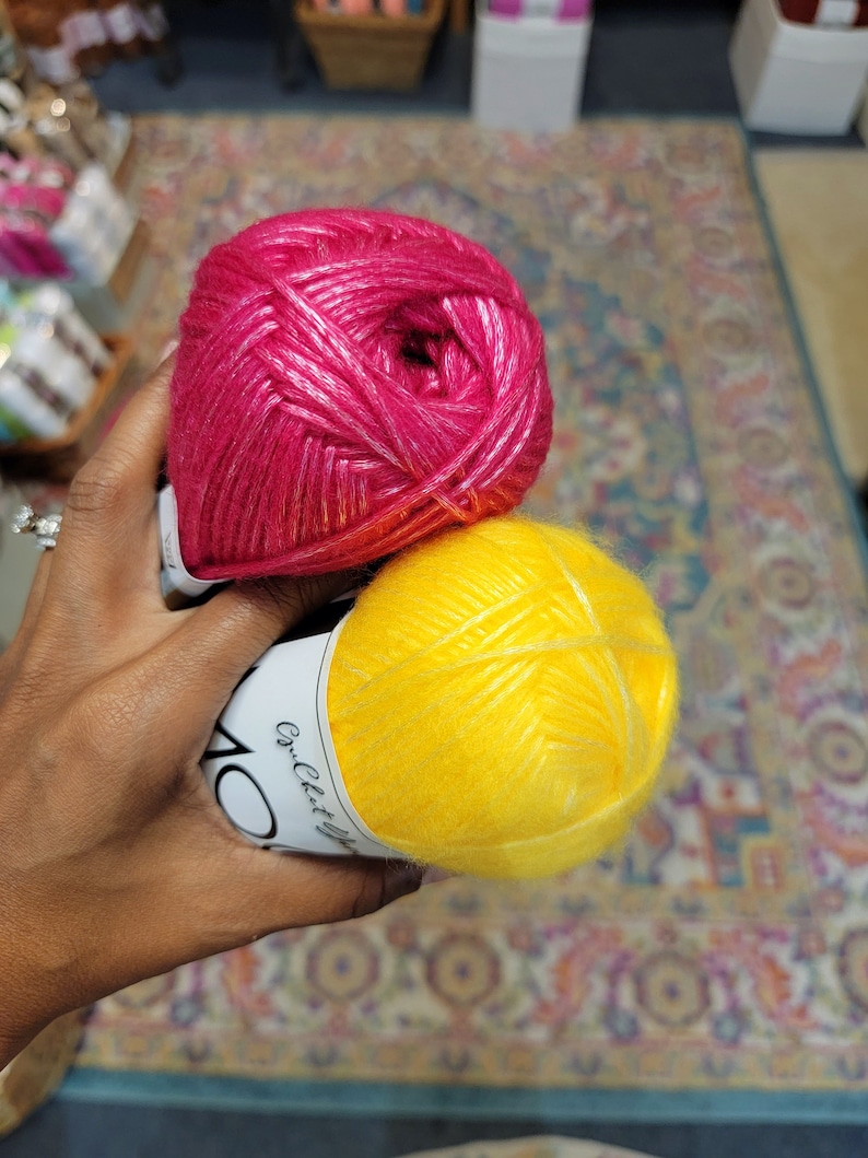 May include: Two skeins of yarn, one in a vibrant magenta pink and the other in a bright sunshine yellow. Each skein features a white paper label with the text Crochet Up, showcasing a soft, fuzzy texture with a subtle sheen throughout the fibers.
