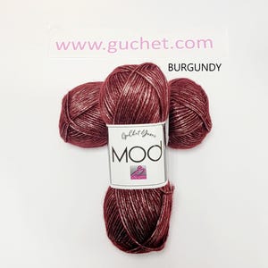 May include: Three skeins of burgundy yarn with a metallic sheen, arranged in a cluster. A white paper label wraps around the center, featuring the text "Guchet Yarns MOD" and a small logo. The text "www.guchet.com" and "BURGUNDY" appear above.