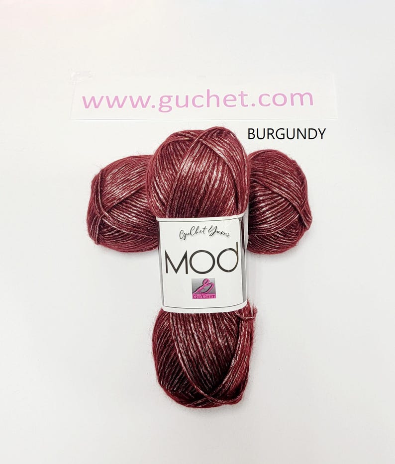 May include: Three skeins of burgundy yarn with a metallic sheen, arranged in a cluster. A white paper label wraps around the center, featuring the text "Guchet Yarns MOD" and a small logo. The text "www.guchet.com" and "BURGUNDY" appear above.